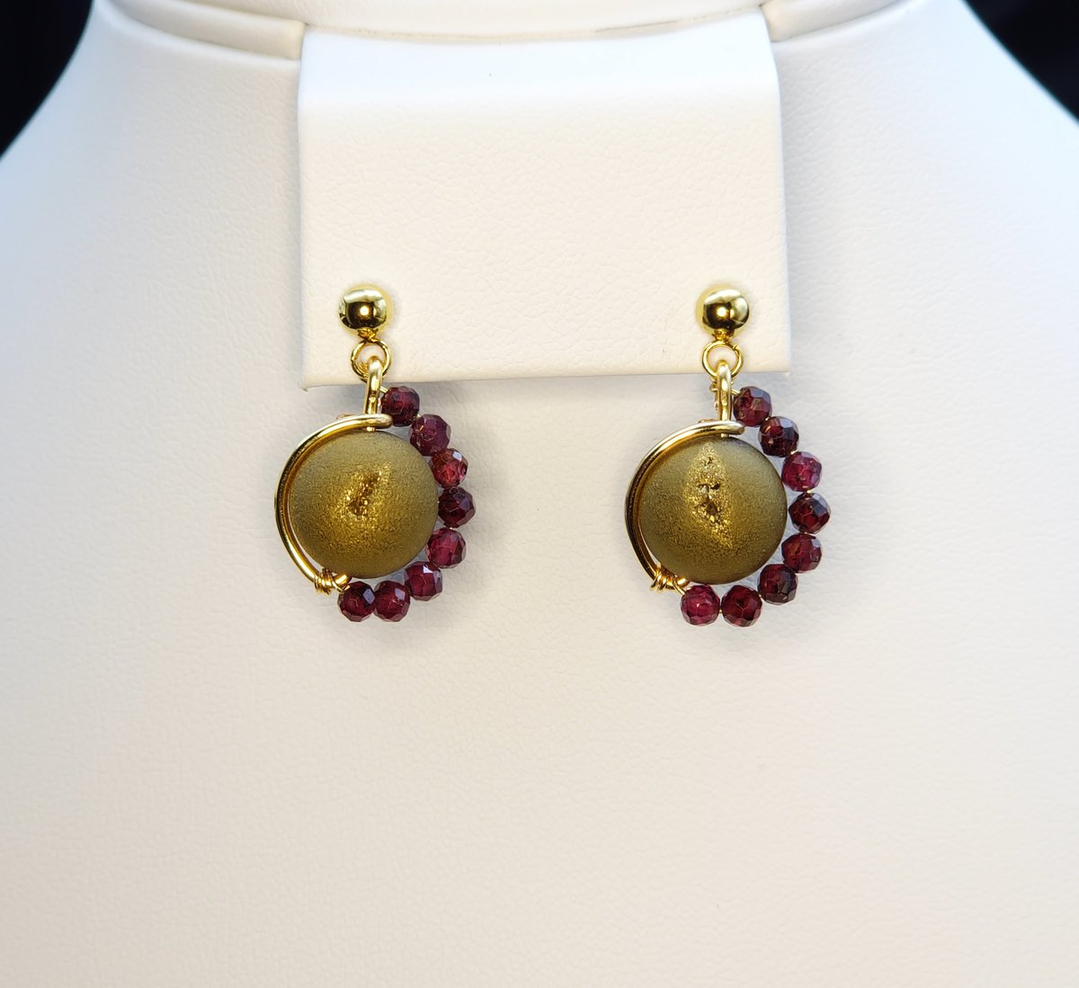An asymmetrictal design featuring a green/gold Druzy Agate center, garnet accents and stainless steel gold earring post