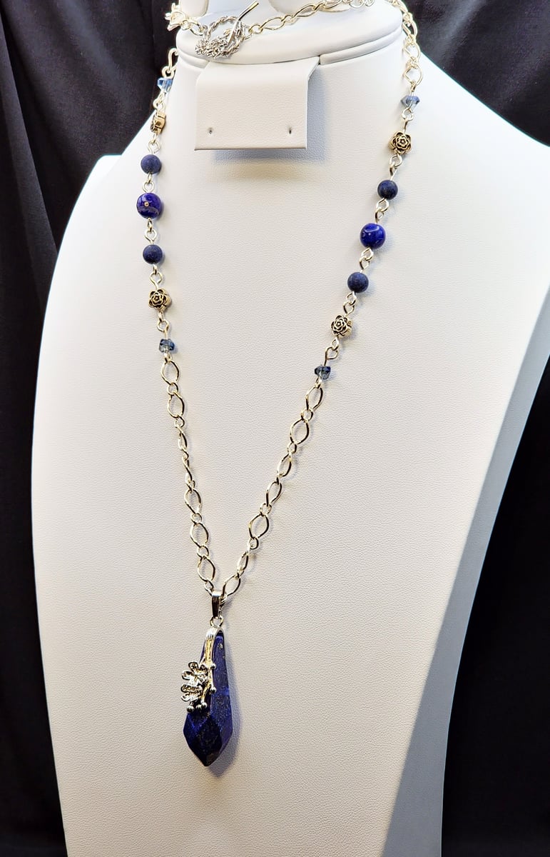 A large Lapis Lazuli pendant accompanied by silver figaro chain, matte Lapis Lazuli and glass beads. A platinum plated brass figural flower toggle clasp brings it together