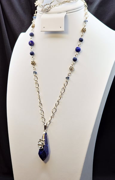 A large Lapis Lazuli pendant accompanied by silver figaro chain, matte Lapis Lazuli and glass beads. A platinum plated brass figural flower toggle clasp brings it together